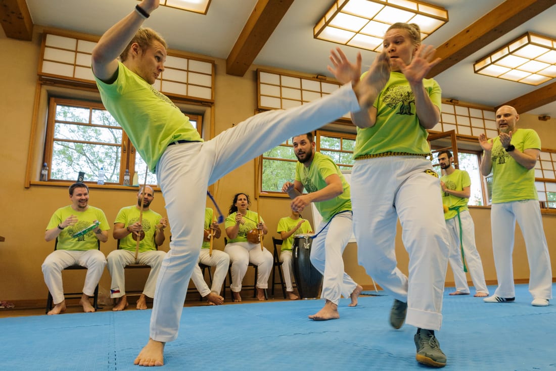 Dva capoeiristé při tréninku v sále, jeden vysoko kopá, ostatní hrají na nástroje v kruhu